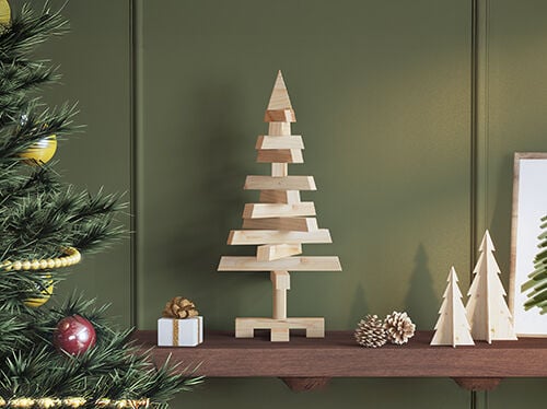 A wooden shelf with small Christmas decorations including a modern wooden tree in the center, two smaller wooden trees, pinecones, and a small wrapped gift, with part of a decorated Christmas tree visible on the left side.