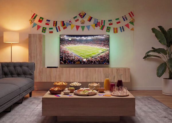 Living room prepared for a football match viewing, with a TV showing a crowded stadium, a sofa facing a wooden coffee table filled with snacks and drinks, and colorful international flags decorating the wall.
