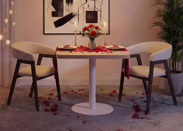 Romantic dining setup in a modern living room with a small round white table and two upholstered chairs, set with plates, candles, a vase of red roses, and scattered rose petals on the table and rug, framed by soft curtain lights, abstract wall art, and a green indoor plant for a cozy Valentine&rsquo;s Day atmosphere.