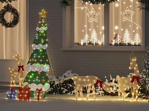 An outdoor Christmas display with illuminated reindeer figures wearing red bows, a green and white lighted tree with colorful bulbs, and wrapped gift decorations in the snow, set in front of a house with glowing window decorations of stars and trees.