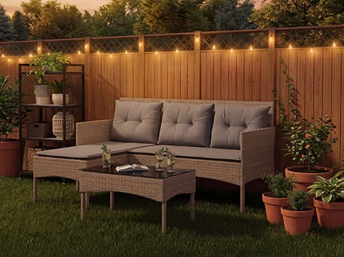 Rattan garden lounge set with grey cushions including a three-seat sofa, footstool and coffee table on a lawn with string lights and potted plants