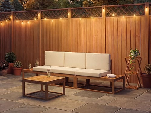 Three-seat modular outdoor sofa with cream cushions and a matching low coffee table, set on a stone patio against a warm wooden wall