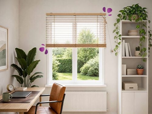 A home office corner with a wood Venetian blind installed above a large window, softly filtering natural light and overlooking a green garden outside. A wooden desk and leather chair sit below, with a tall plant and white open shelving with small cacti visible on either side, creating a calm and airy workspace.