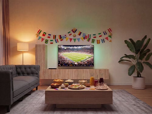 Living room set up for watching a football match, with a TV showing a stadium game, a sofa facing a wooden coffee table filled with snacks and drinks, and colorful international flag garlands decorating the wall. Click to see the products in the collection.
