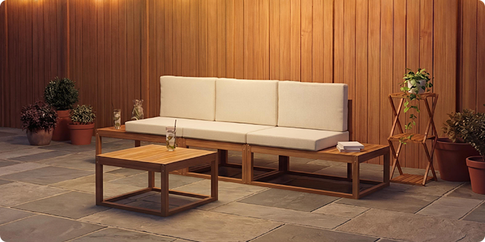 Three-seat modular outdoor sofa with cream cushions and a matching low coffee table, set on a stone patio against a warm wooden wall